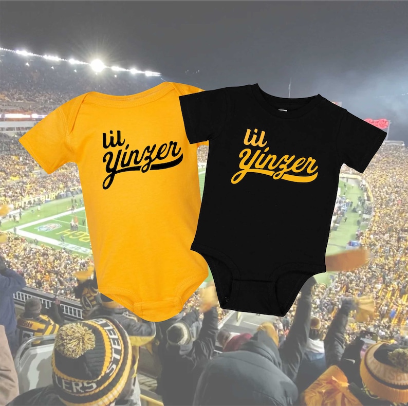 K&ouml;nnte beinhalten: Zwei Baby-Bodys, einer gelb und einer schwarz, mit dem Text "lil' yinzer" in einer Retro-Schrift. Die Bodys befinden sich vor einem verschwommenen Hintergrund einer Stadionmenge.