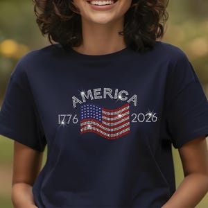 Smiling woman wearing navy t-shirt with rhinestone USA flag and America 250th anniversary design outdoors.