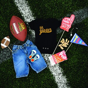 K&ouml;nnte beinhalten: Ein schwarzer Body mit dem Text "lil' yinzer" in goldenen Buchstaben, ein brauner Football, eine blaue Jeans, ein Paar blaue und wei&szlig;e Turnschuhe, ein roter und wei&szlig;er "You're No. 1" Schaumfinger, eine T&uuml;te Erdn&uuml;sse, eine T&uuml;te Popcorn und eine blau-wei&szlig;e "Team"-Flagge.
