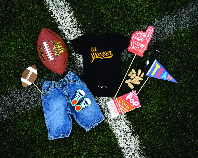 K&ouml;nnte beinhalten: Ein schwarzer Body mit dem Text "lil' yinzer" in goldenen Buchstaben, ein brauner Football, eine blaue Jeans, ein Paar blaue und wei&szlig;e Turnschuhe, ein roter und wei&szlig;er "You're No. 1" Schaumfinger, eine T&uuml;te Erdn&uuml;sse, eine T&uuml;te Popcorn und eine blau-wei&szlig;e "Team"-Flagge.