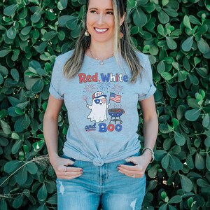 May include: A light gray t-shirt with the text "Red, White and Boo" and a cartoon ghost wearing sunglasses and a USA hat, holding a sparkler and American flag. The ghost is next to a barbecue grill.