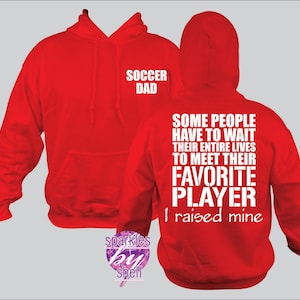May include: Red hooded sweatshirt with the text "Soccer Dad" on the front and "Some people have to wait their entire lives to meet their favorite player I raised mine" on the back.