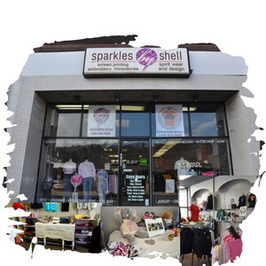 K&ouml;nnte beinhalten: Ein Ladenlokal mit einem Schild, auf dem "sparkles by shell screen printing embroidery rhinestones spirit wear and design" steht. Das Ladenlokal hat eine gro&szlig;e Schaufensterauslage mit Kleidung, Accessoires und anderen Artikeln zum Verkauf. Der Laden befindet sich in der 246 W Main Street und die Telefonnummer lautet (724) 510-3990.
