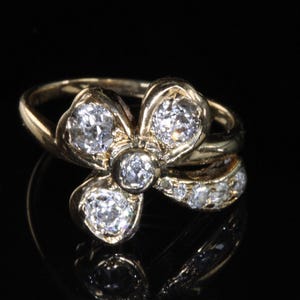 Puede incluir: Anillo de oro con dise&ntilde;o floral, con diamantes redondos y transparentes. La banda del anillo tambi&eacute;n tiene diamantes m&aacute;s peque&ntilde;os, sobre un fondo negro.