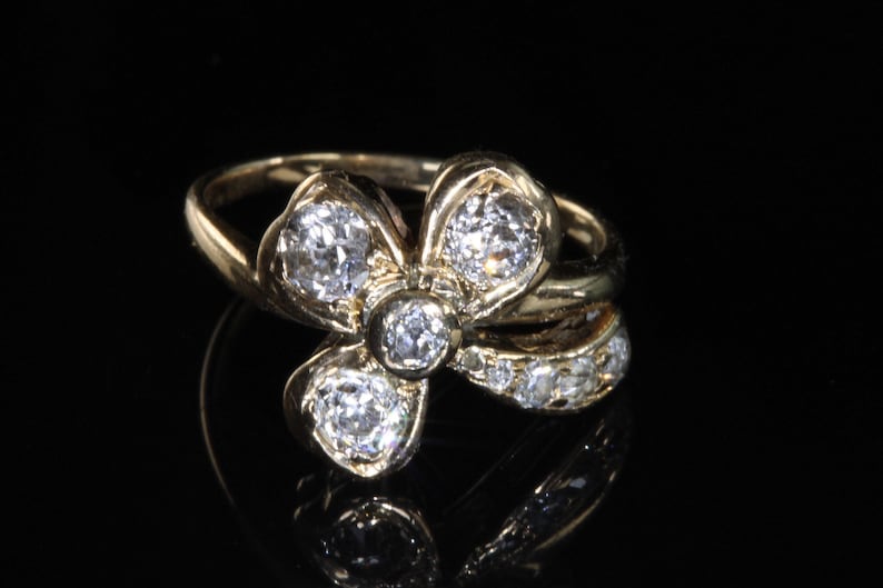 Puede incluir: Anillo de oro con dise&ntilde;o floral, con diamantes redondos y transparentes. La banda del anillo tambi&eacute;n tiene diamantes m&aacute;s peque&ntilde;os, sobre un fondo negro.