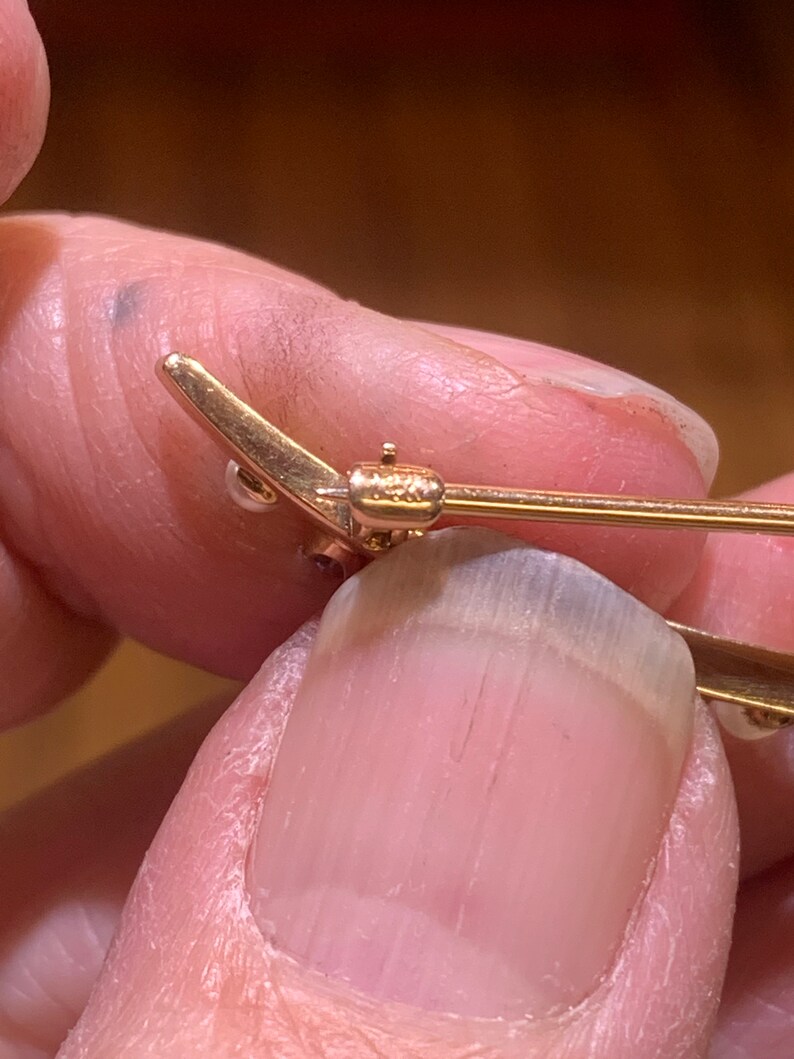 May include: Close-up of a gold-toned brooch with a pearl and a red stone. The brooch has a safety pin closure.