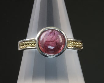 Könnte beinhalten: Ein silberner Ring mit einem rosafarbenen Edelstein, der in einer goldenen Fassung sitzt. Die Ringschiene ist mit einem geflochtenen Golddrahtmuster verziert.