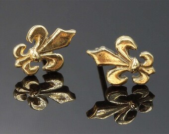 Könnte beinhalten: Ein Paar goldfarbene Fleur-de-Lis-Ohrstecker. Die Ohrringe haben die Form der stilisierten Lilienblume mit drei Blütenblättern und einem zentralen Element. Die Ohrringe werden auf einer reflektierenden Oberfläche präsentiert.