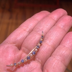 May include: A crescent moon shaped brooch with alternating purple gemstones and white pearls. The brooch is made of gold metal.