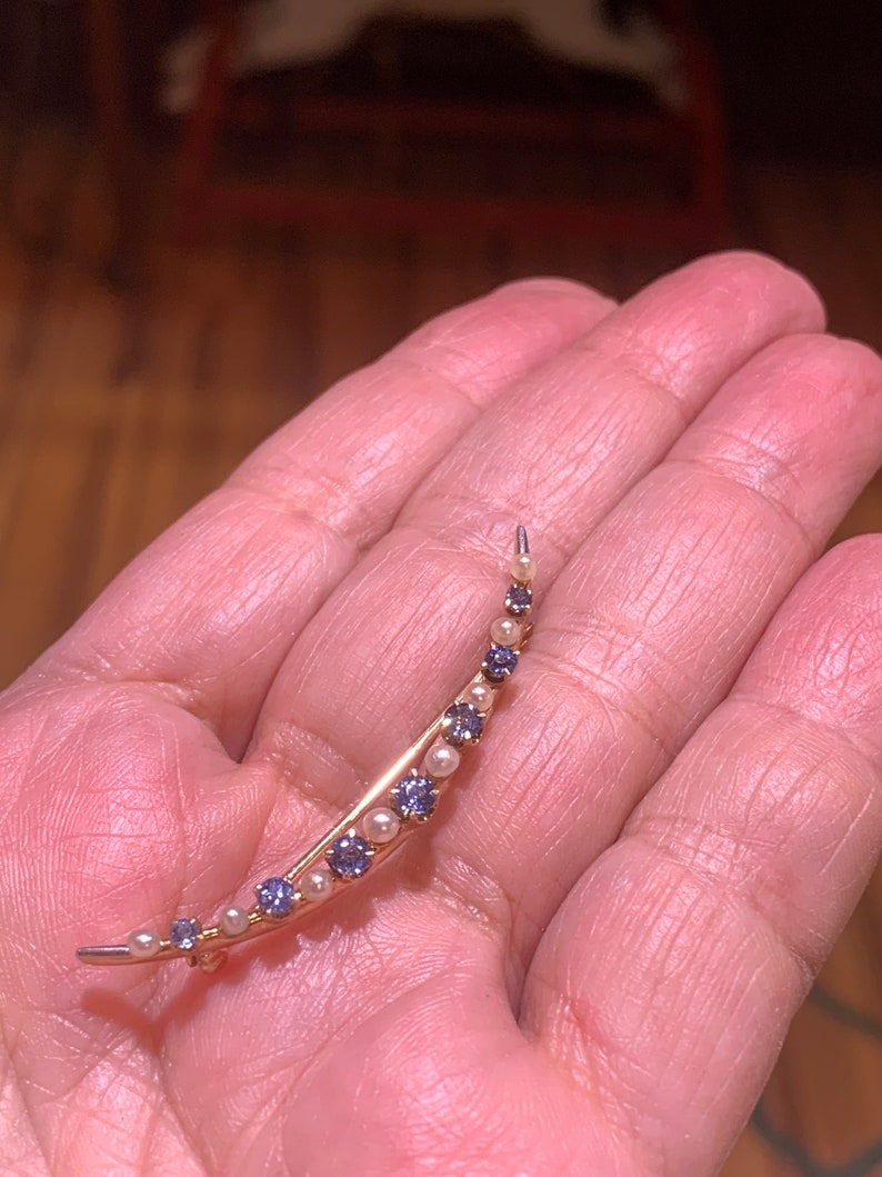 May include: A crescent moon shaped brooch with alternating purple gemstones and white pearls. The brooch is made of gold metal.