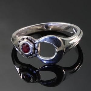 Puede incluir: Un anillo de plata con una piedra roja engastada en una forma hexagonal. El anillo tiene forma de llave inglesa.