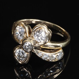 Puede incluir: Un anillo de oro con un dise&ntilde;o floral, con m&uacute;ltiples piedras preciosas transparentes. La banda del anillo es lisa y las piedras preciosas est&aacute;n engastadas en un grupo, creando un efecto brillante. El anillo se muestra sobre un fondo negro.