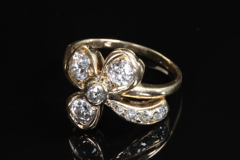 Puede incluir: Un anillo de oro con un dise&ntilde;o floral, con m&uacute;ltiples piedras preciosas transparentes. La banda del anillo es lisa y las piedras preciosas est&aacute;n engastadas en un grupo, creando un efecto brillante. El anillo se muestra sobre un fondo negro.