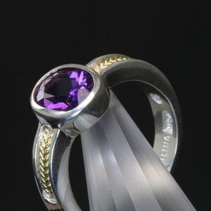 May include: A silver ring with a purple amethyst gemstone set in a bezel setting. The band is accented with a thin band of gold.