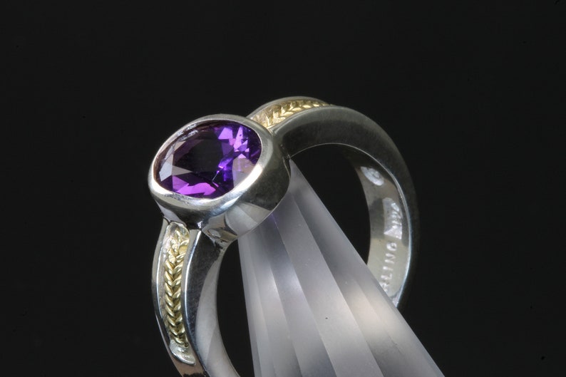 May include: A silver ring with a purple amethyst gemstone set in a bezel setting. The band is accented with a thin band of gold.