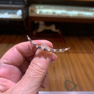 May include: A crescent moon shaped brooch with a white stone in the center and a row of small blue and white stones around the edge.