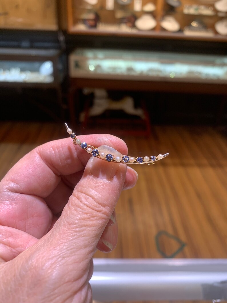 May include: A crescent moon shaped brooch with a white stone in the center and a row of small blue and white stones around the edge.