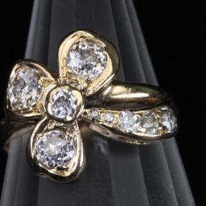 Puede incluir: Anillo dorado con dise&ntilde;o floral y cinco piedras preciosas claras. Piedras m&aacute;s peque&ntilde;as adornan la banda. El anillo se muestra sobre un fondo oscuro, resaltando sus intrincados detalles.