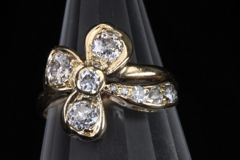 Puede incluir: Anillo dorado con dise&ntilde;o floral y cinco piedras preciosas claras. Piedras m&aacute;s peque&ntilde;as adornan la banda. El anillo se muestra sobre un fondo oscuro, resaltando sus intrincados detalles.