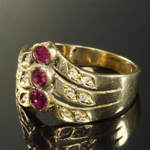 Op de afbeelding: Een gouden ring met drie rode edelstenen en een rij kleine diamanten op elke band.