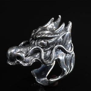 May include: A silver dragon head ring. The ring features a detailed dragon head design with intricate scales, horns, and facial features. The ring is made of a shiny, metallic material and is set against a black background.