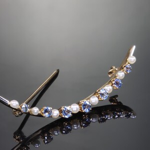 May include: A gold crescent-shaped brooch with blue and white stones. The brooch has a pin on the back for attaching to clothing.