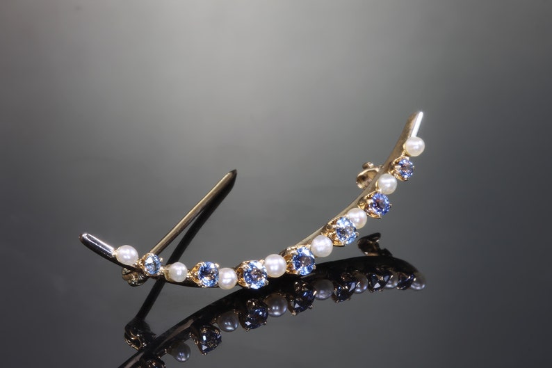 May include: A gold crescent-shaped brooch with blue and white stones. The brooch has a pin on the back for attaching to clothing.