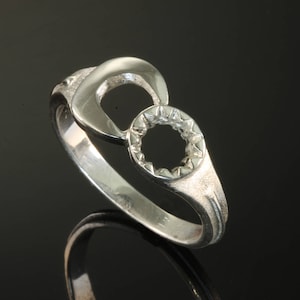 Puede incluir: Un anillo de plata con un diseño único que presenta dos círculos entrelazados, uno con un recorte circular y el otro con un patrón de estrella.