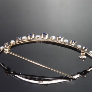 May include: A gold brooch with a curved design, set with small blue sapphires and white pearls. The brooch has a pin and catch closure.