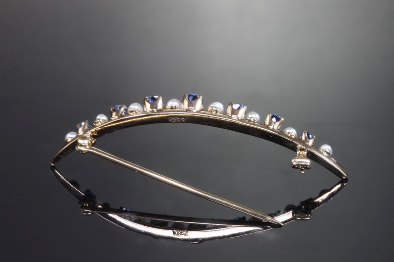 May include: A gold brooch with a curved design, set with small blue sapphires and white pearls. The brooch has a pin and catch closure.