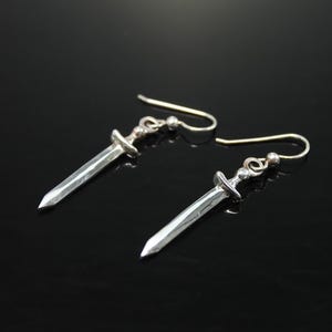 Pode incluir: Um par de brincos de espada de prata com um design simples. Os brincos são feitos de metal e têm um acabamento prateado.