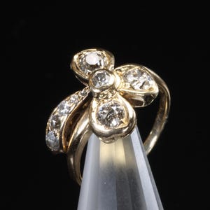 Puede incluir: Un anillo dorado con dise&ntilde;o floral, engastado con m&uacute;ltiples piedras preciosas transparentes. El anillo se muestra sobre un fondo negro, resaltando sus intrincados detalles y brillo. Una joya.