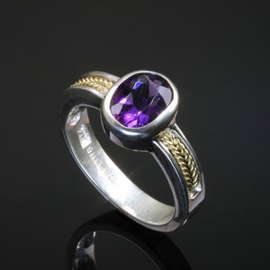 May include: A silver ring with a purple amethyst gemstone set in a gold bezel. The band is decorated with a braided gold wire design.