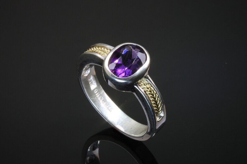 May include: A silver ring with a purple amethyst gemstone set in a gold bezel. The band is decorated with a braided gold wire design.