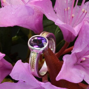 May include: A silver and gold ring with an oval amethyst gemstone set in a bezel setting. The ring is surrounded by pink flowers.