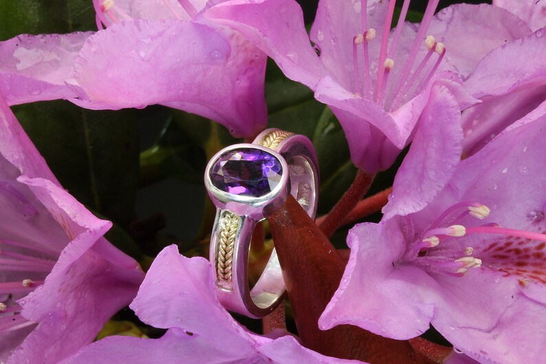 May include: A silver and gold ring with an oval amethyst gemstone set in a bezel setting. The ring is surrounded by pink flowers.
