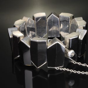 Puede incluir: Un brazalete de plata hecho de formas geométricas, con una cadena adjunta. El brazalete está hecho de muchas piezas pequeñas y rectangulares que están conectadas entre sí.