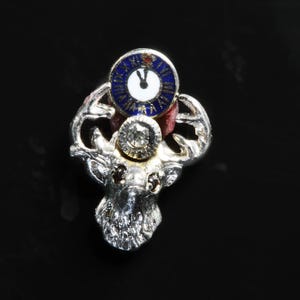 May include: A silver pin with a detailed deer head design. The deer's antlers are adorned with a blue and white enamel clock face and a clear diamond-like stone.