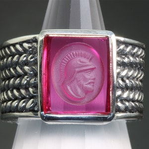 Puede incluir: Un anillo de plata con una piedra cuadrada rosa con una imagen tallada de una persona con un casco. La banda del anillo está trenzada.