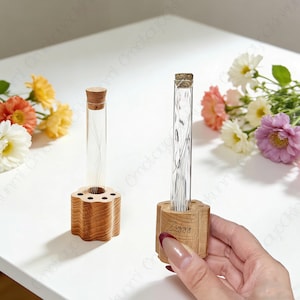 May include: Two clear glass test tubes with cork stoppers, each containing a delicate arrangement, sit in wooden holders. One holder is engraved with the name "Ziggy". The holders are on a white surface, with colorful flowers in the background.