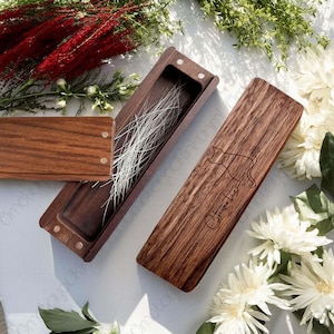 May include: A dark wooden pencil case with a carved cat design and the name "Charlie". The case is open, showing a compartment with white strands. The case has a magnetic closure and is surrounded by flowers.