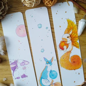 Peut inclure: Trois marque-pages illustrés représentant différents personnages d'animaux. Le premier marque-page représente un lapin blanc avec des accents violets tenant une fleur rose. Le deuxième marque-page représente un chat bleu et blanc avec une étoile sur le front tenant un orbe bleu. Le troisième marque-page représente un lapin blanc avec des ailes orange tenant une pêche.