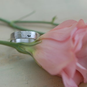 Può includere: Un anello in argento martellato con un piccolo cuore inciso sulla fascia. L'anello è appoggiato su un fiore rosa.