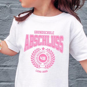 Op de afbeelding: Een wit t-shirt met roze tekst "Grundschule Abschluss" en een cirkelvormig embleem. Het embleem bevat het nummer "4B" en de naam "Lena 2026". Het t-shirt wordt door een kind gedragen.