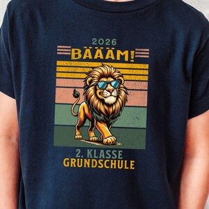 Könnte beinhalten: Ein dunkelblaues T-Shirt mit einer Grafik eines Löwen, der eine blaue Sonnenbrille trägt. Das Design enthält horizontale Streifen in Gelb, Orange und Grün sowie den Text 2026 BÄÄÄM! und 2. KLASSE GRUNDSCHULE in Gelb und Weiß.