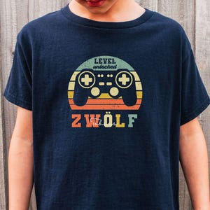 Op de afbeelding: Marineblauwe t-shirt met een retro videogamecontroller-afbeelding. Het ontwerp bevat de tekst "LEVEL unlocked" boven de controller en het nummer "ZWÖLF" eronder, in een vintage kleurenschema.