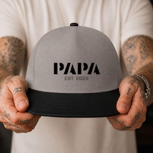 Könnte beinhalten: Eine graue Snapback-Kappe mit schwarzem Schirm und einem schwarzen Knopf oben. Auf der Vorderseite der Kappe befindet sich der Schriftzug "PAPA" in einer fetten, stilisierten schwarzen Schrift, darunter "EST. 2020" in kleinerer Schrift.