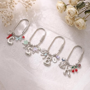 May include: Four silver-toned oval carabiner keychains, each featuring a large letter charm and several smaller decorative charms. The letters include C, S, 8, and K, accompanied by various colorful enamel and crystal accents like butterflies, cherries, and flowers.
