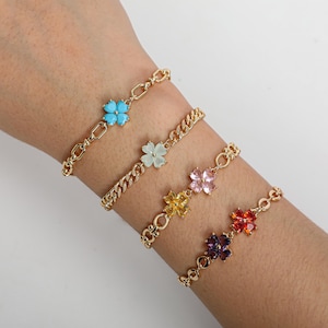 以下が含まれることがあります： それぞれ異なる色の四つ葉のクローバーチャームが付いた、4つのゴールドトーンのブレスレット。 クローバーチャームは、ターコイズ、ライトグリーン、ピンク、イエロー、オレンジ、パープルの色合いです。 ブレスレットは手首にあります。