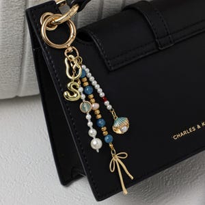 May include: Black handbag with a gold-tone charm. The charm features a gold letter "S", beads in various colors, and a seashell pendant. The handbag has a gold clasp and the brand name "CHARLES & K" is printed on the side.